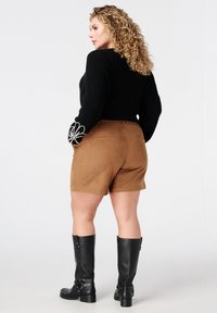 Krullendharige vrouw in een zwarte trui met witte bloeiende manchetten, bruine shorts en zwarte knielange laarzen, staand en zijwaarts kijkend.