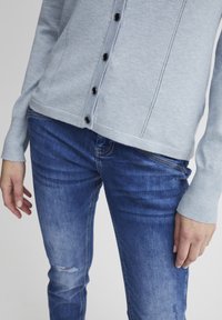 Gaiši zils adīts kardigāns ar melnām pogām un redzamām šuvēm, kombinēts ar nolietotām zilām džinsām, kurām ir plīstoša detaļa pie ceļa.