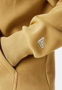 Manica di un sweat shirt giallo con polsino a costine e un piccolo logo bianco ricamato. Texture liscia e design casual.