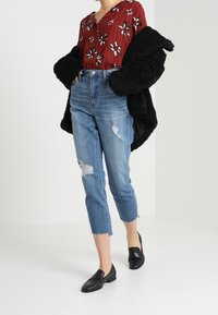 Blusa a fiori rossi con dettagli a bottoni, abbinata a jeans chiari strappati e mocassini neri. Cappotto nero peloso drappeggiato sulle spalle.
