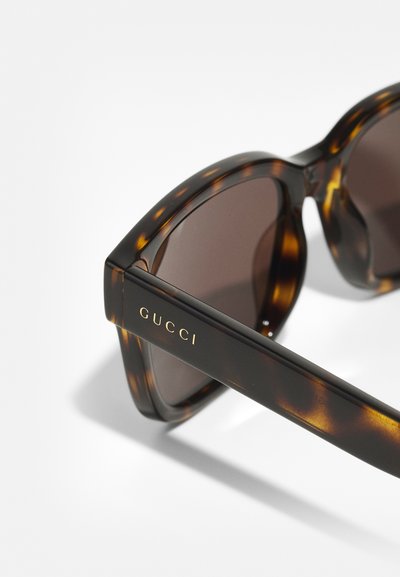 Tortoiseshell sluneční brýle s hnědými skly, mají silný rám, jemný vzor a zlaté logo "GUCCI" na nožičce.