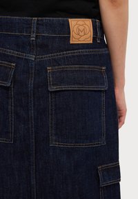 Denimrok van donkerblauwe stof met contrasterende gele stiksels, twee achterzakken en een bruine leren label met een ingedrukt logo.