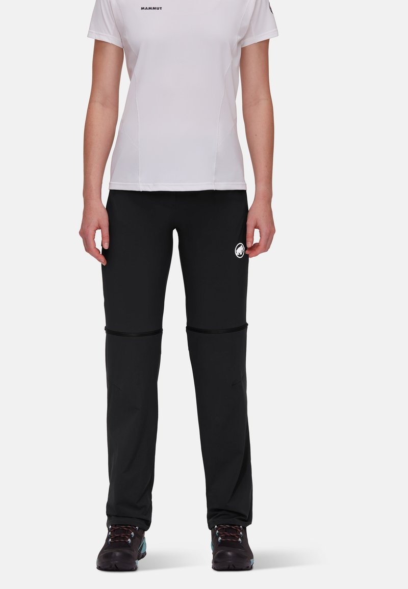 Persoon met een wit Mammut T-shirt met korte mouwen en zwarte outdoorbroek met ritssluiting, en wandelschoenen, staande tegen een witte achtergrond.