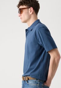 Πόλο μπλουζάκι κοντομάνικο σε σκούρο μπλε βαμβάκι. Διαθέτει κλασικό γιακά, δύο κουμπιά στον λαιμό και χαλαρή εφαρμογή. Φορεμένο με ανοιχτό μπλε τζιν.