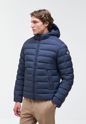 Giovane uomo che indossa una giacca a vento blu navy con cappuccio, in piedi con una mano nella tasca dei pantaloni beige, guarda verso sinistra.