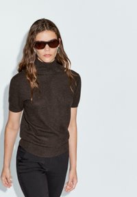 Brun kortärmad polokrage tröja med en mjuk textur och figurnära design, kombinerad med svarta byxor och mörka solglasögon.