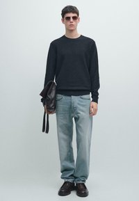 Maglione nero texturizzato, jeans azzurri e scarpe di pelle marrone. Tiene in mano una borsa di pelle marrone. Occhiali da sole marroni completano il look.