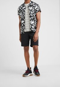 Man med svart och vit blommig kortärmad skjorta över en grårandig t-shirt, svarta shorts och svarta sneakers med röda snören.