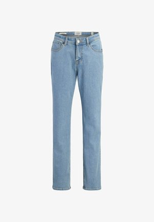 Garçon debout, la main sur la hanche, portant un t-shirt noir, un jean bleu clair et des chaussures marron à lacets, souriant et regardant sur le côté.