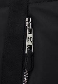 Zwarte ritssluiting met een zilveren trekhaak met een 'K' logo, tegen een gestructureerde stofachtergrond.