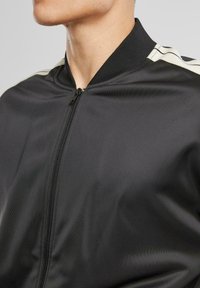 Veste bomber légère noire en tissu lisse. Dotée d'une fermeture éclair à l'avant et d'un col côtelé, avec des rayures contrastantes sur les épaules.