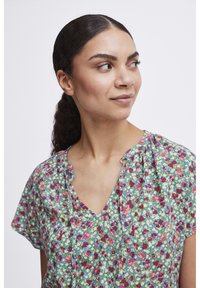 Blouse fleurie avec un fond vert ornée de fleurs rouges et bleues, à manches courtes, décolleté en V, avec des détails plissés sur le devant.