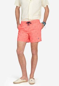 Bañador de color coral con cintura ajustable, que presenta un pequeño patrón y logo en blanco. Combinado con una camisa de color claro con cuello y zapatos tipo slip-on en beige.