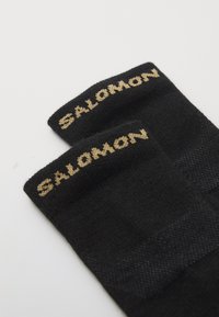 Meias desportivas pretas com textura canelada, logótipo dourado "SALOMON" e áreas reforçadas para maior durabilidade.