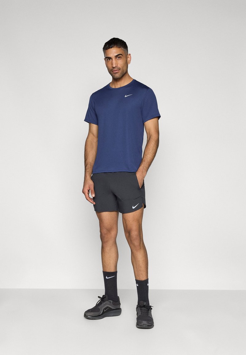 Marineblaues sportliches T-Shirt mit kurzen Ärmeln, kombiniert mit schwarzen Shorts mit Seitentaschen, schwarzen Socken und schwarzen Sportschuhen.