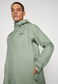 Chaqueta impermeable verde claro con cremallera frontal, capucha y detalle de logo. Presenta una textura suave y bolsillos laterales.