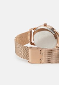 Bracelet de montre en maille rose doré avec une boucle en métal poli, présentant un boîtier rond lisse et un fond en verre clair. Détails de surface texturée visibles.