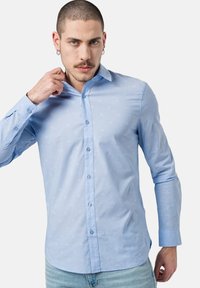 Camisa de botón azul claro con una sutil textura de estampado. Presenta un cuello estándar y mangas largas, confeccionada en una tela suave.