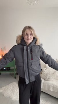 Νεαρή γυναίκα μέσα σε εσωτερικό χώρο, φορώντας γκρι χειμερινό παλτό με κουκούλα από γούνα, στέκεται με τα χέρια ελαφρώς ανοιχτά, χαμογελώντας προς την κάμερα.