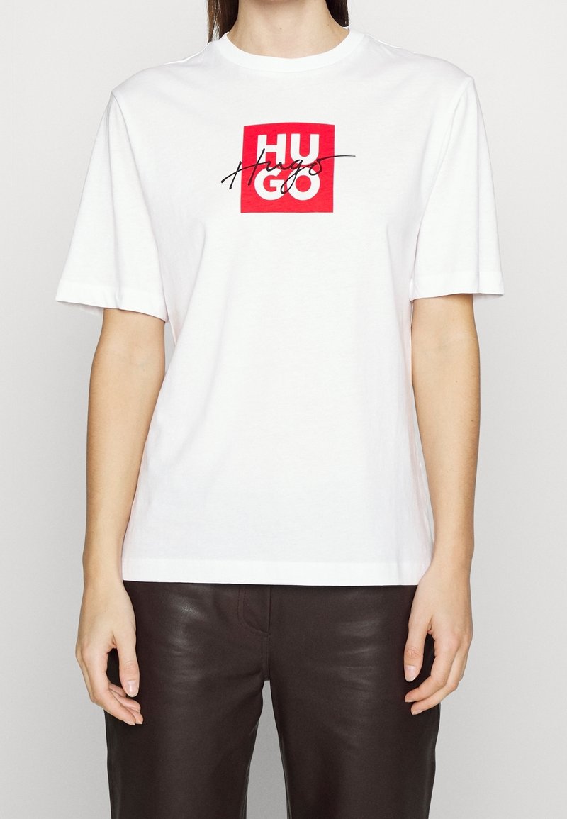 Camiseta de algodón blanca de manga corta, presenta un diseño de logo en forma de cuadrado rojo con la palabra "HUGO" en letras negras gruesas centrada en el pecho.