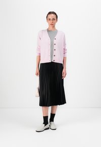 Cardigan de color rosa claro con cierre de botones, camisa gris debajo, falda negra con tablas y zapatillas blancas. Incluye un pequeño bolso rosa.