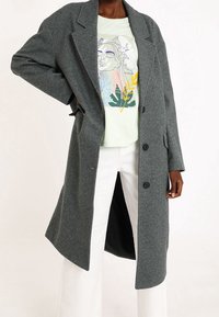 Cappotto grigio a spina di pesce con design monopetto, grandi risvolti e tre bottoni. Indossato sopra una t-shirt grafica verde chiaro e pantaloni bianchi.