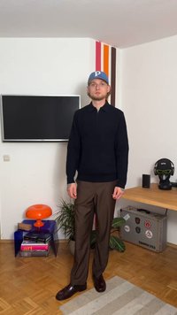 Νεαρός άνδρας με μπλε καπέλο, μαύρη ζακέτα, καφέ παντελόνι και σκούρα παπούτσια στέκεται σε ένα δωμάτιο με τηλεόραση, πορτοκαλί λάμπα, φυτά και γραφείο.