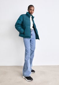 Chaqueta de plumas en color verde azulado con cuello alto y diseño acolchado, combinada con jeans acampanados de color azul claro y zapatillas negras. Estilo simple y funcional.