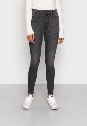 Jeans negros slim-fit con cintura alta, que presentan un sutil desvanecimiento y un cierre de botón clásico, combinados con zapatillas blancas.