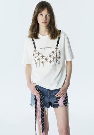 Joven mujer con camiseta blanca de gran tamaño con arnés decorativo y shorts vaqueros con detalles de tela a rayas, posando con las manos en los bolsillos.