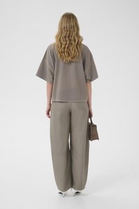 Άτομο με μακριά κυματιστά ξανθά μαλλιά, φοράει χαλαρή τούνελ τοπ χρώματος τόουπ και παντελόνια με φαρδιές μπάντες, κρατώντας μια μικρή τσάντα, στέκεται σε λευκό φόντο.