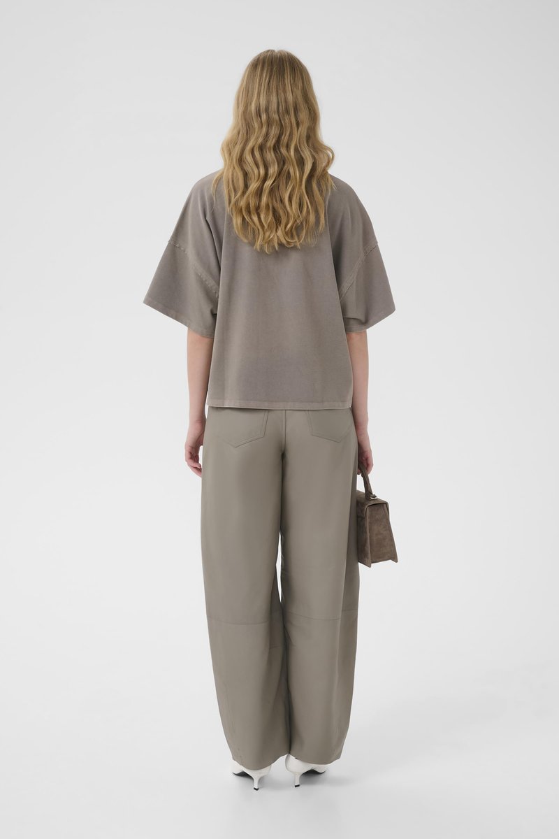 Personne aux longs cheveux blonds ondulés portant un haut ample taupe et un pantalon à jambes larges, tenant un petit sac, debout sur un fond blanc.