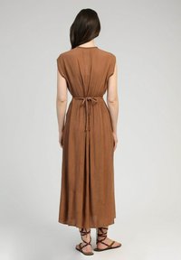 Robe longue sans manches marron avec un dos froncé et une taille à nouer. Fabriquée en tissu doux et texturé atteignant le mollet. Sandales avec des lanières croisées.