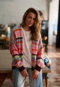 Mladá žena s vlnitými vlasy, která má na sobě barevně pruhovaný cardigan a bílý tričko, opírá se o kulečníkový stůl v rozmazané místnosti.