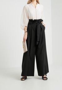 Blouse rose clair avec un col et des manches bouffantes, associée à un pantalon large noir taille haute avec un nœud. Tenue d'une pochette beige.
