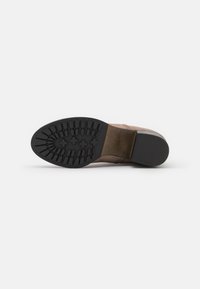 Bottines en daim marron vues par le dessous, présentant une semelle en caoutchouc noir avec des motifs de relief et un talon empilé sur un fond blanc.