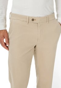 Pantaloni in cotone beige con taglio dritto. Presenta un cinturino con bottoni e tasche laterali. Texture liscia, senza motivi o accenti.