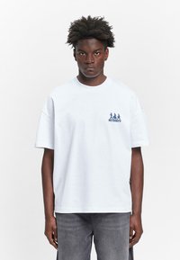 Weißes Baumwoll-T-Shirt mit kurzen Ärmeln, lockerer Passform, mit einem kleinen blauen Grafik-Design und Text im linken Brustbereich.