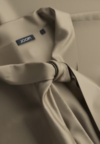 Beige JOOP!-Mantel mit einem gebundenen Gürtel, wobei der Kragenbereich und das Markenlabel im Inneren des Kleidungsstücks im Fokus stehen.