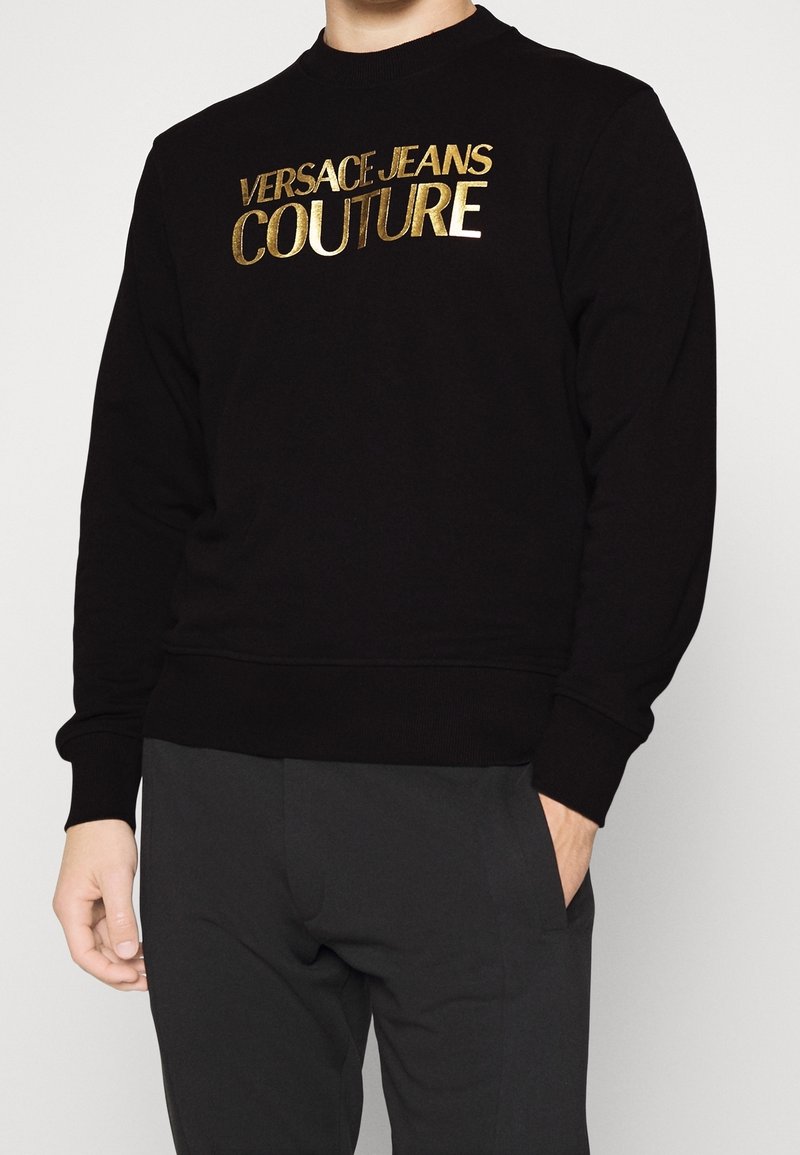 Homme portant un sweat-shirt noir avec des lettres dorées "Versace Jeans Couture" sur la poitrine et un pantalon noir, une main dans la poche.