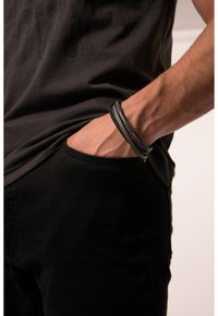 Set di bracciali in pelle a tre filamenti, con texture intrecciate e lisce, in tonalità nere, indossati su un polso vicino a una tasca di pantaloni neri.