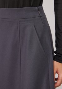 Jupe grise sur mesure avec une texture lisse, dotée de poches latérales et d'une fermeture éclair dissimulée à l'arrière pour une finition soignée.