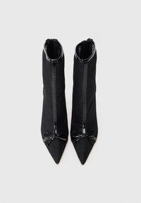 Μαύρες γυναικείες ankle boots με ψηλό τακούνι, φτιαγμένες από υλικό με υφή πλέγματος, μυτερή μύτη, λεπτομέρειες από λουστρίνι και διακοσμητική λεπτομέρεια από φιόγκο.