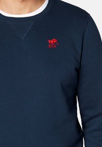 Granatowy sweter z ribbowym okrągłym dekoltem. Na piersi znajduje się małe czerwone haftowane logo konia oraz napis "POLO SYLT".