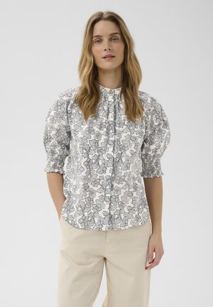 Femme aux longs cheveux portant un chemisier blanc à motifs floraux noirs et manches bouffantes, associé à un pantalon beige clair sur un fond uni.