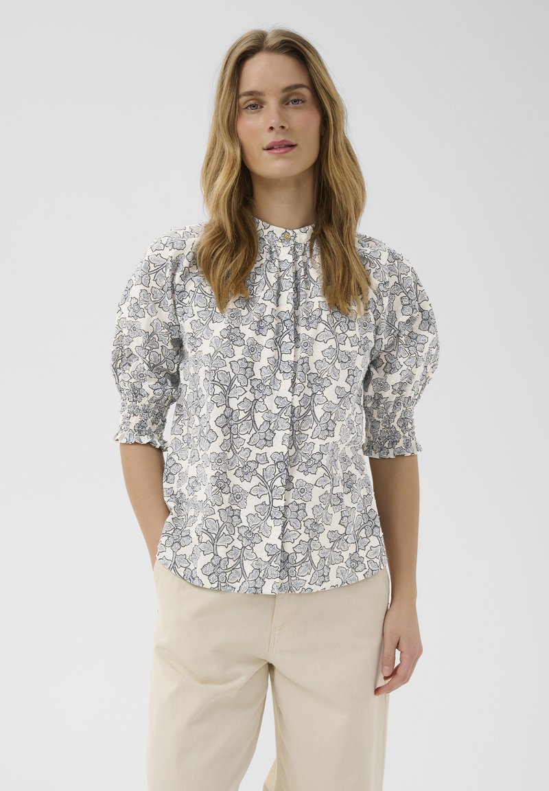 Femme aux longs cheveux portant un chemisier blanc à motifs floraux noirs et manches bouffantes, associé à un pantalon beige clair sur un fond uni.