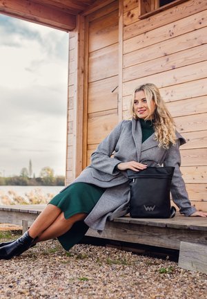 Grau-wollener Mantel über einem grünen Rippkleid, kombiniert mit einer schwarzen Lederhandtasche mit Logo. Sie trägt schwarze Stiefeletten und sitzt auf einem Holzdeck.