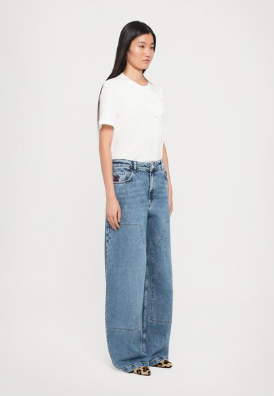 White short-sleeve t-shirt with textured detailing, paired with wide-leg blue denim jeans featuring patches and a waistband label, worn with patterned shoes.