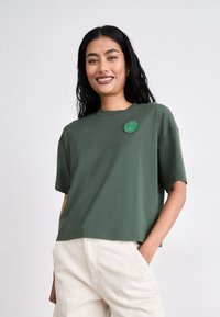 Grønn bomulls t-skjorte med rund hals og korte ermer. Har en rund, grønn brodert lapp på brystet. Enkelt design, avslappet passform.