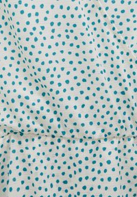 Tissu blanc avec de petits pois bleu sarcelle irréguliers et une texture côtelée horizontale subtile, légèrement plié et froncé au niveau d'une couture.